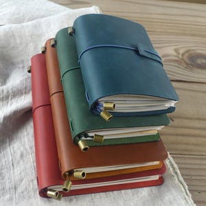 May include: A stack of leather-bound journals in various colors: red, brown, green, and teal. Each journal has a matching elastic closure and brass-colored metal accents. The journals are stacked on a light-colored fabric and wooden surface.