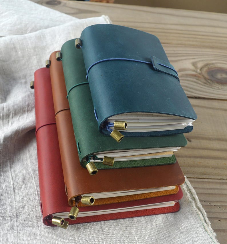 May include: A stack of leather-bound journals in various colors: red, brown, green, and teal. Each journal has a matching elastic closure and brass-colored metal accents. The journals are stacked on a light-colored fabric and wooden surface.