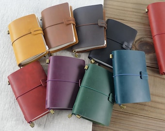 May include: A collection of leather-bound journals in various colours including yellow, brown, dark brown, black, red, purple, green, and blue. Each notebook features a wrap-around elastic closure, a metal fastener, and visible paper pages.