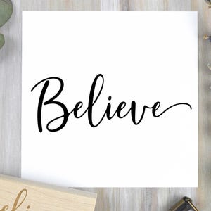 May include: A white square print with the word "Believe" in elegant black cursive script. The print is set against a light wood grain background, with sprigs of lavender and eucalyptus leaves adding a natural touch.