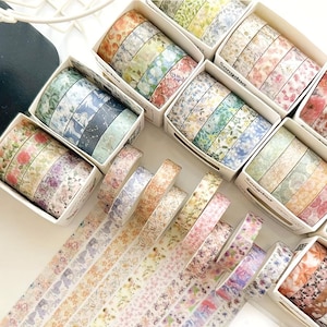 May include: A collection of decorative washi tape rolls featuring various floral, botanical, and butterfly patterns in soft pastel and vibrant colors. The rolls are organized in white cardboard boxes, with several individual rolls unspooled to display.