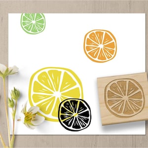 May include: A collection of citrus-themed art and a wooden stamp. The art features illustrations of lemon, lime, and orange slices in yellow, green, orange, and black. The wooden stamp has a carved orange slice design.