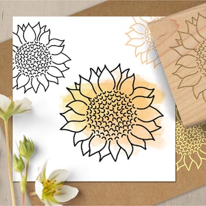 May include: A collection of sunflower-themed art and a wooden stamp. The art features black line drawings of sunflowers, some with yellow watercolor backgrounds. The wooden stamp has a sunflower design carved into its surface. The items are arranged on a white background.