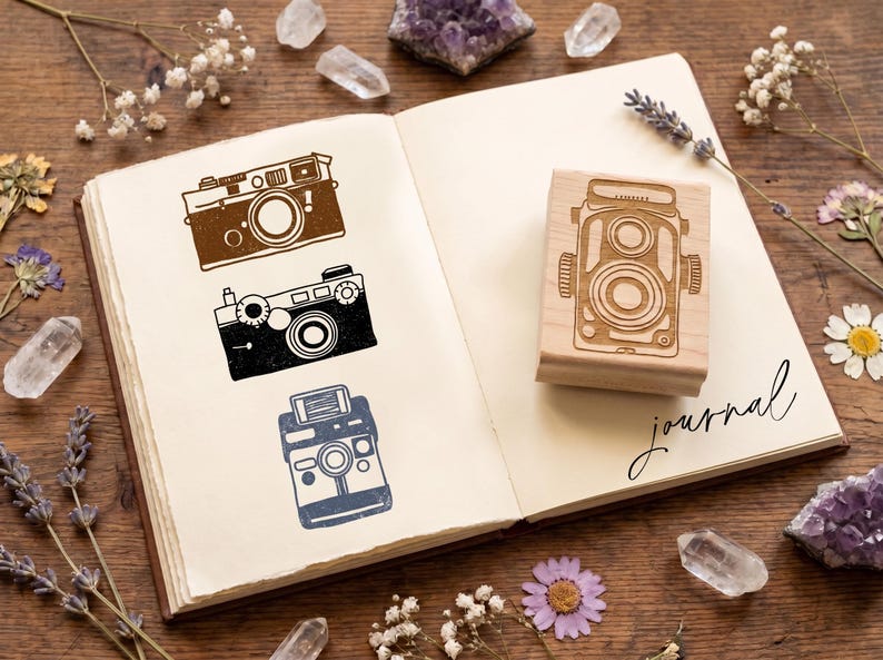May include: An open journal displays camera illustrations in brown, black, and blue. A wooden camera stamp is on the right page. The word "journal" is written in cursive. The background is a wooden surface. The journal is surrounded by crystals.