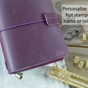 May include: A purple leather journal with a matching closure strap and elastic band. The journal is personalized with the name "Alex" and the text "Personalize with hot stamped name or initials". Gold letter stamps are visible.