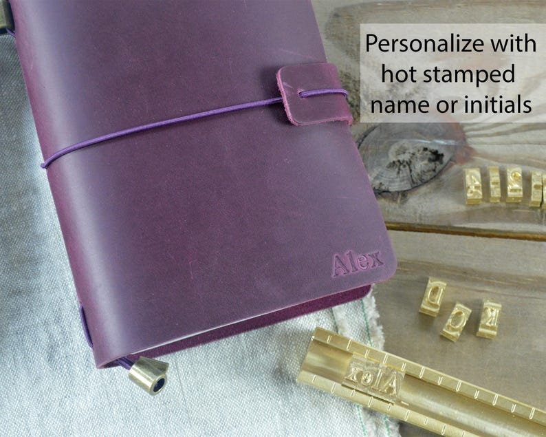 May include: A purple leather journal with a matching closure strap and elastic band. The journal is personalized with the name "Alex" and the text "Personalize with hot stamped name or initials". Gold letter stamps are visible.