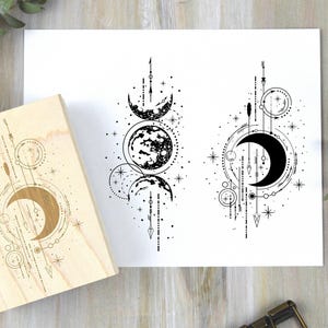 May include: A wooden stamp with a crescent moon design and a detailed celestial illustration on white paper. The artwork features moons, stars, and arrows in a black and white color scheme. The stamp is made of light-colored wood.