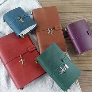 May include: A collection of leather journals in various colors: red, brown, green, blue, and purple. Each journal is secured with a strap and features a decorative charm, such as a key, airplane, or dragonfly. The journals are arranged on a wooden surface.