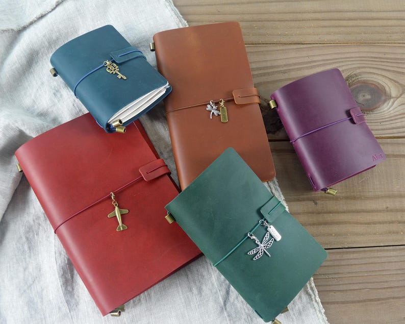 May include: A collection of leather journals in various colors: red, brown, green, blue, and purple. Each journal is secured with a strap and features a decorative charm, such as a key, airplane, or dragonfly. The journals are arranged on a wooden surface.