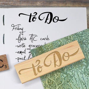 May include: A wooden stamp with the words "To Do" in a cursive font, next to a notepad with a handwritten to-do list. A Scrabble tile with the letter "C" and the number "3" is also present. The notepad has a list that includes "finish ATC cards".
