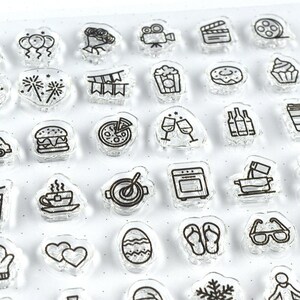 May include: Clear acrylic stamps with black line art designs. The stamps feature various icons, including a cake, balloons, a film reel, popcorn, a burger, pizza, wine glasses, and a coffee cup. These stamps are ideal for crafting and scrapbooking projects.