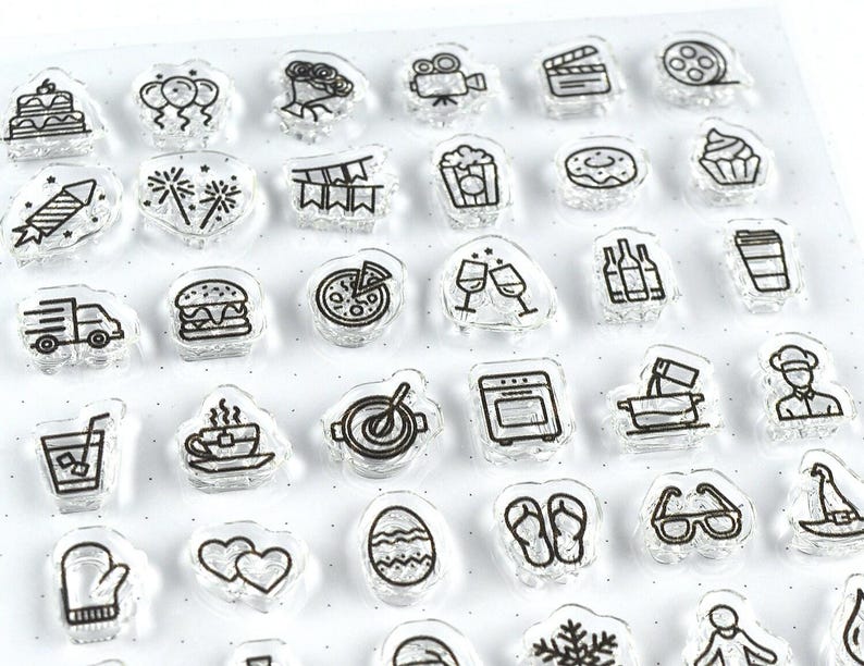 May include: Clear acrylic stamps with black line art designs. The stamps feature various icons, including a cake, balloons, a film reel, popcorn, a burger, pizza, wine glasses, and a coffee cup. These stamps are ideal for crafting and scrapbooking projects.