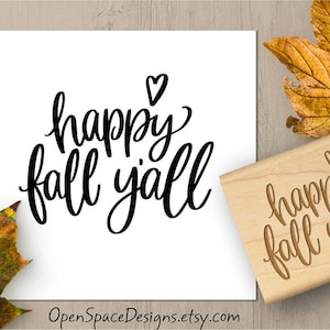 May include: A white card with the phrase "happy fall y'all" in black script, with a heart above the word "happy". A wooden stamp with the same text is next to the card. Autumn leaves are in the background.