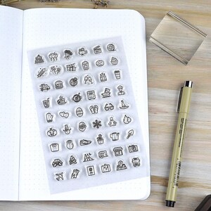 May include: A clear sheet of stamps with various small black line art icons, including food, travel, and holiday-themed images. The sheet is next to an open dot grid notebook, a clear stamp block, and a pen on a wooden surface.
