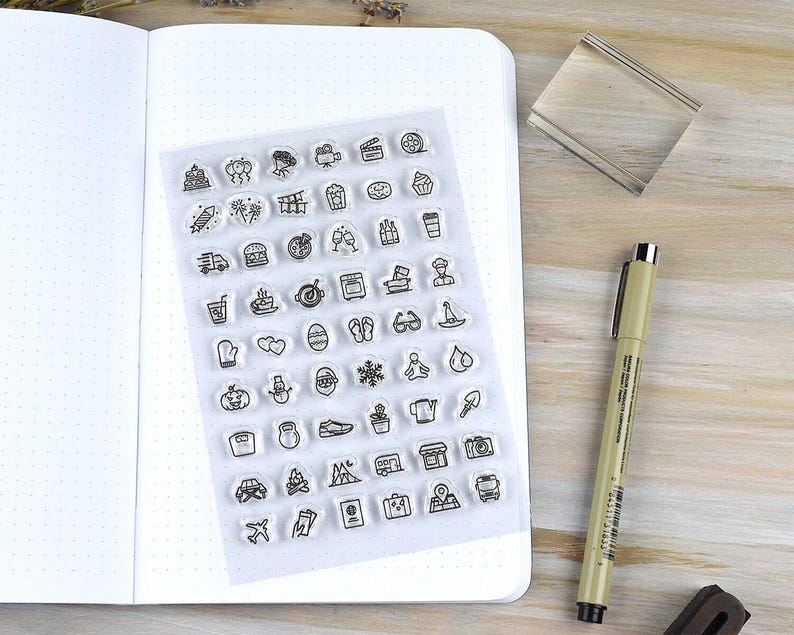 May include: A clear sheet of stamps with various small black line art icons, including food, travel, and holiday-themed images. The sheet is next to an open dot grid notebook, a clear stamp block, and a pen on a wooden surface.