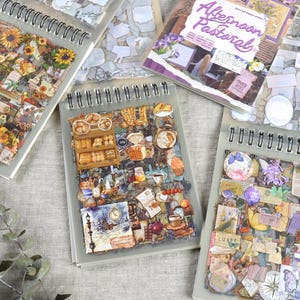 May include: Several spiral-bound sticker books with decorative sticker sheets. One book features sunflowers and vintage elements, another showcases bakery items, and a third displays purple floral and butterfly designs. The top book reads "Afternoon Pastoral".