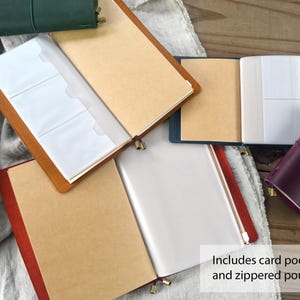 May include: Leather journals in various colors, including green, brown, and burgundy, are displayed open to reveal card pockets and a zippered pouch. The journals have a classic design with a leather cover and a closure.