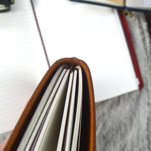 May include: Close-up of a brown leather journal with cream-colored lined pages. The journal is open, revealing the interior pages and the binding. Other journals are visible in the background.