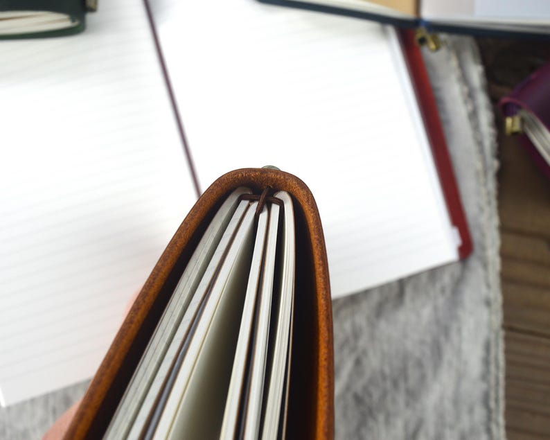 May include: Close-up of a brown leather journal with cream-colored lined pages. The journal is open, revealing the interior pages and the binding. Other journals are visible in the background.