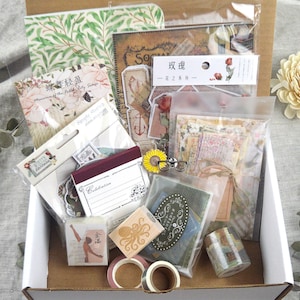 May include: A curated box filled with stationery supplies. Includes decorative paper, stickers, washi tape, and stamps. The items feature floral, vintage, and botanical designs. A sunflower and small glasses are also included.