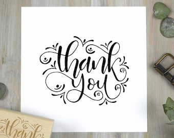 May include: A white card with the words "thank you" in elegant black script. The text is surrounded by decorative flourishes. A wooden stamp with the word "thank" is visible in the lower left corner.