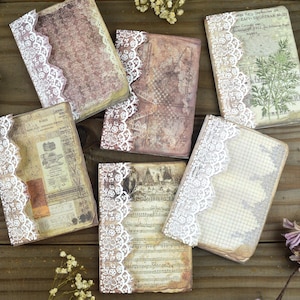 May include: A collection of vintage-style journals with aged paper pages and decorative white lace trim. The journals feature various printed designs and textures, creating a rustic and artistic aesthetic. The journals are arranged on a wooden surface.