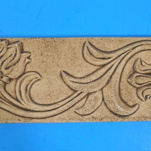 May include: A rectangular tan ceramic tile featuring a raised relief design of two stylized flowers connected by a flowing vine. The surface has a textured, matte finish with darker brown shading within the recessed areas of the floral pattern.