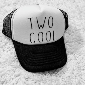 May include: A black and white trucker hat with the words "TWO COOL" in black lettering. The hat has a white front panel, a black brim, and a black mesh back.