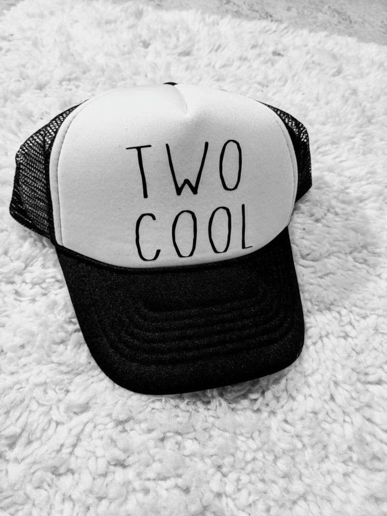 May include: A black and white trucker hat with the words "TWO COOL" in black lettering. The hat has a white front panel, a black brim, and a black mesh back.
