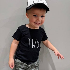 May include: A young child wearing a black t-shirt with the word "TWO" in white, and camouflage pants. The child is also wearing a white and black trucker hat that says "TWO COOL".
