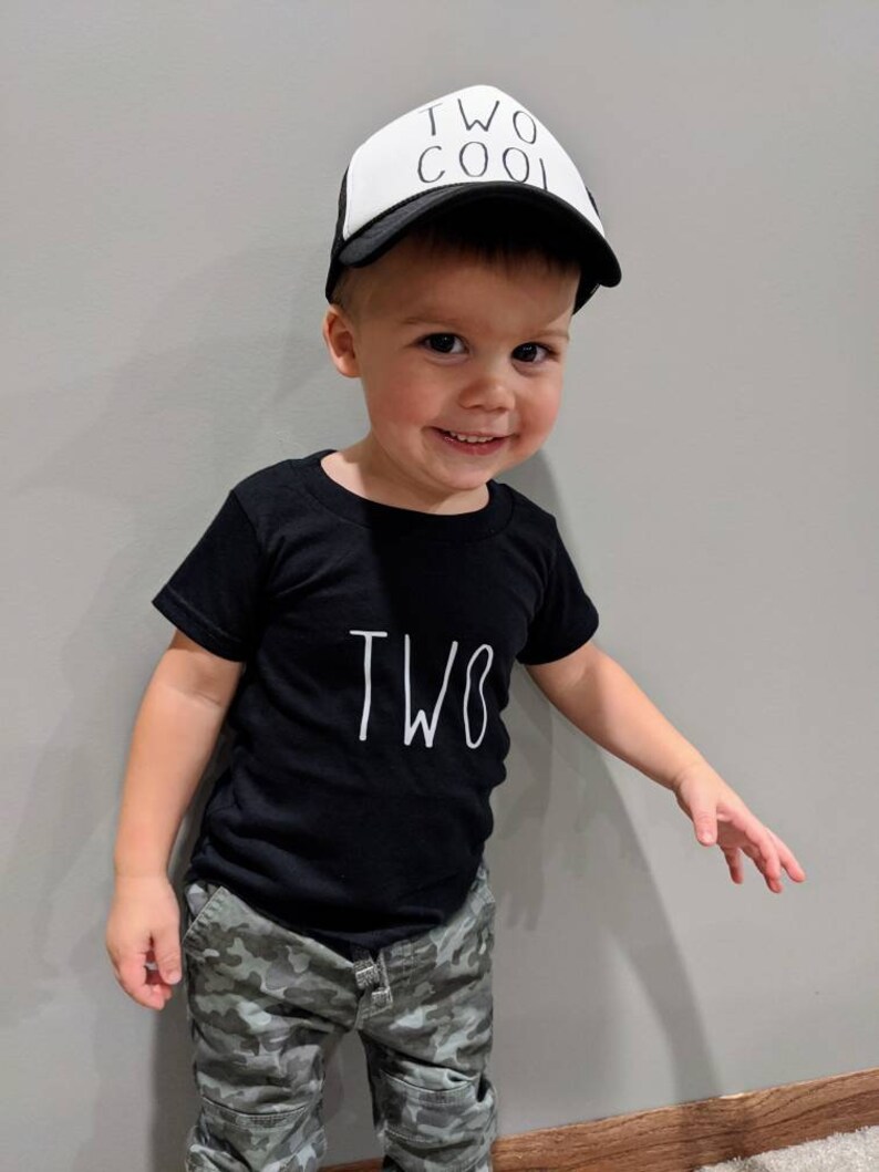 May include: A young child wearing a black t-shirt with the word "TWO" in white, and camouflage pants. The child is also wearing a white and black trucker hat that says "TWO COOL".