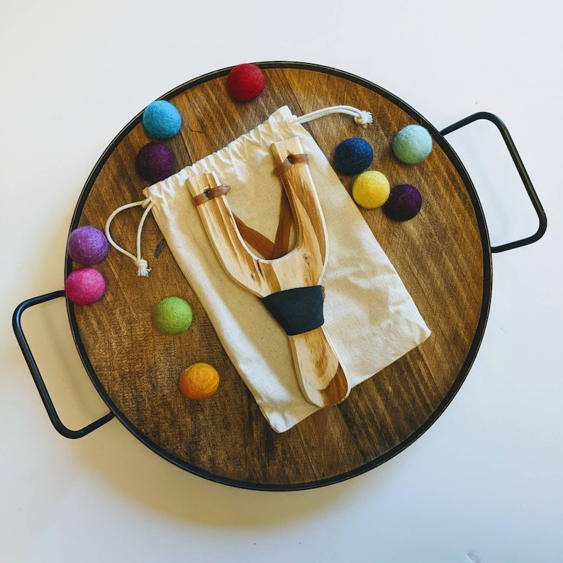 May include: A wooden slingshot with a natural finish, resting on a wooden tray with colorful felt balls.