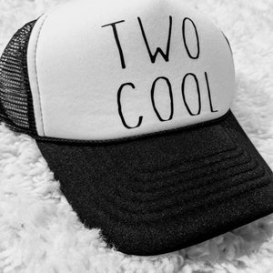 May include: A black and white trucker hat with the words "TWO COOL" printed in black. The hat has a white front panel, a black brim, and a black mesh back.