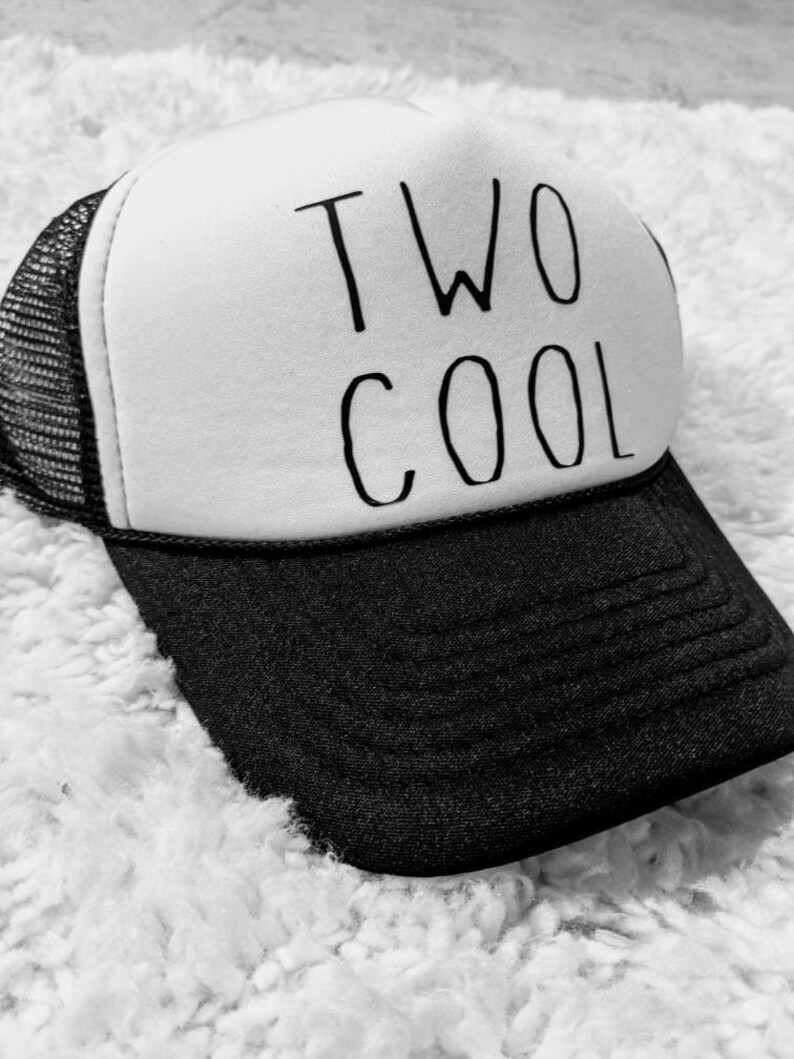May include: A black and white trucker hat with the words "TWO COOL" printed in black. The hat has a white front panel, a black brim, and a black mesh back.