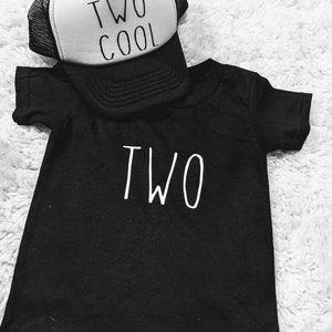 May include: Black toddler t-shirt with the word "TWO" in white. A white and black trucker hat with the words "TWO COOL" is resting on top of the shirt. The items are displayed on a white, fluffy surface.