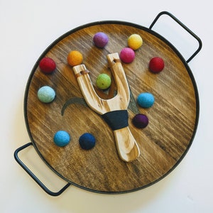May include: A wooden slingshot with a dark blue band and a tray of colorful felt balls. The tray is made of wood and has a dark brown finish.