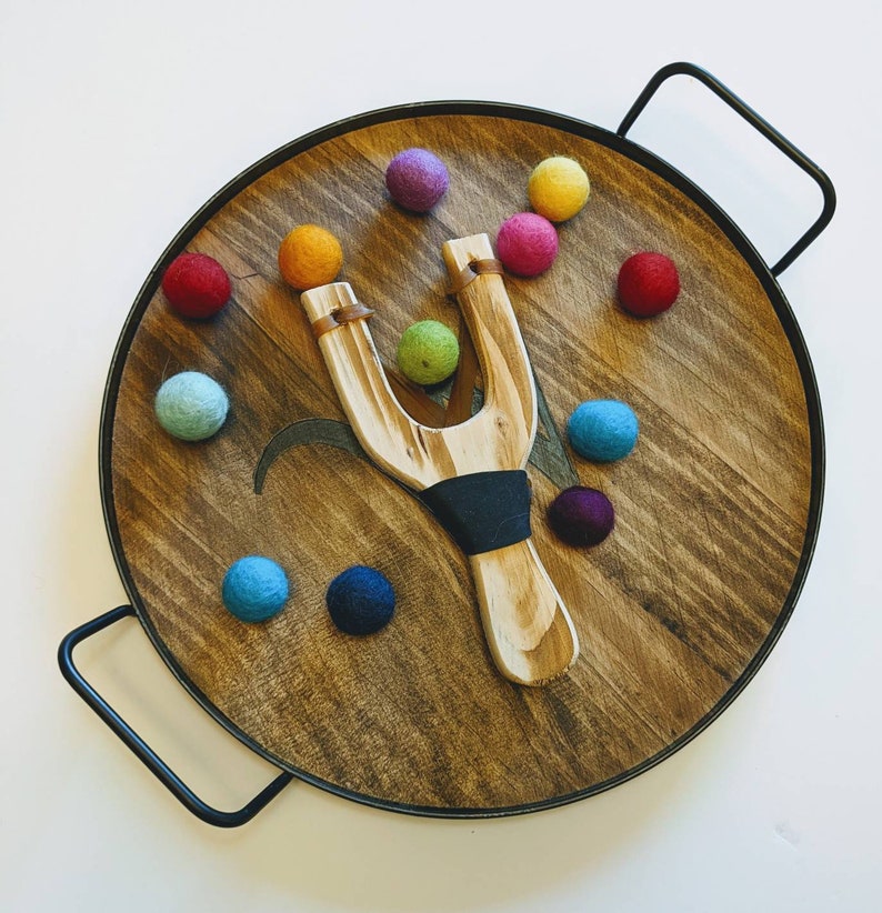 May include: A wooden slingshot with a dark blue band and a tray of colorful felt balls. The tray is made of wood and has a dark brown finish.