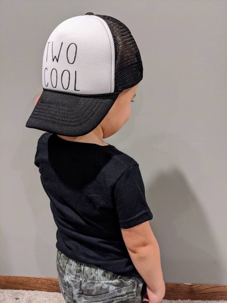 May include: A black and white trucker hat with the words "TWO COOL" printed on the front. The hat has a white front panel, a black brim, and a black mesh back. The hat is worn by a person wearing a black t-shirt.