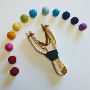May include: A wooden slingshot with a brown leather strap and a black rubber band. The slingshot is surrounded by a rainbow of colorful felt balls.