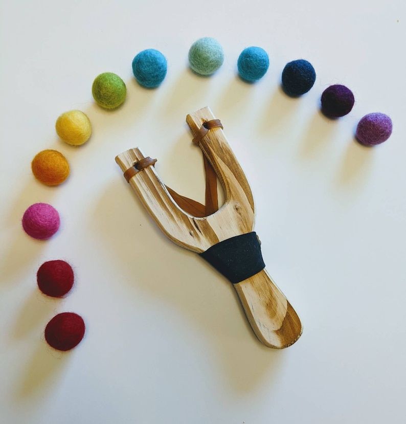 May include: A wooden slingshot with a brown leather strap and a black rubber band. The slingshot is surrounded by a rainbow of colorful felt balls.