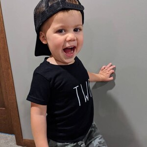 May include: A young child wearing a black mesh trucker hat and a black t-shirt with the word "TWO" printed on it. The child is also wearing camouflage pants and smiling. The child is leaning against a gray wall.