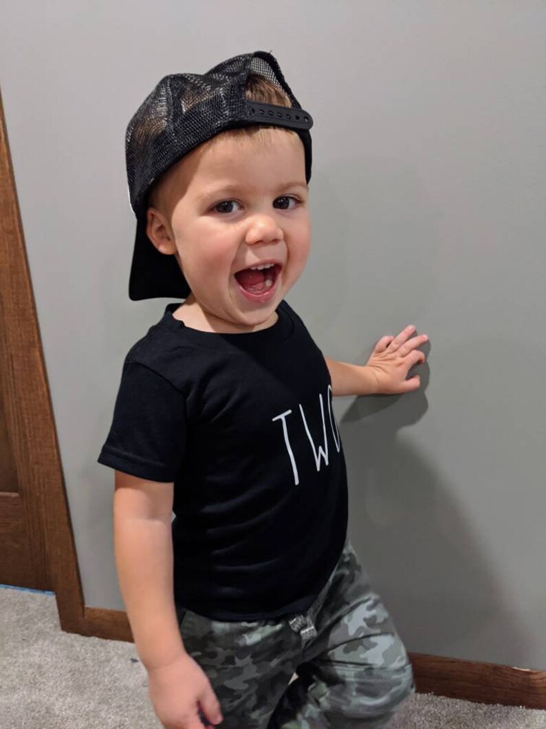 May include: A young child wearing a black mesh trucker hat and a black t-shirt with the word "TWO" printed on it. The child is also wearing camouflage pants and smiling. The child is leaning against a gray wall.