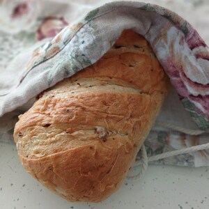 May include: A golden-brown loaf of bread with visible grains and a crusty texture, partially covered by a fabric drawstring bag featuring a floral pattern in shades of pink, green, and beige on a light-colored background.