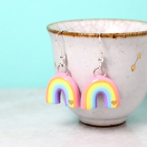A pair of pastel rainbow arch earrings with tiny golden hearts. The earrings are hanging from a small cup in front of a bright blue background.