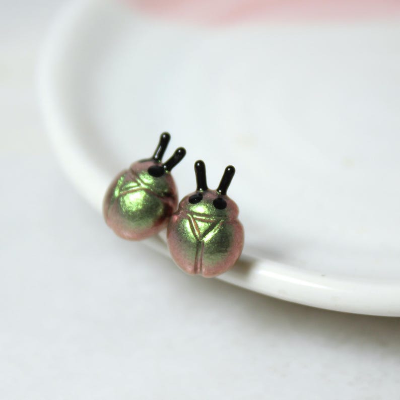 A pair of green and gold beetle stud earrings rest against a white jewelry dish