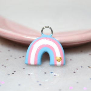 A rainbow arch charm with the colors of the trans pride flag (blue, pink, and white) with a tiny golden heart. The charm is resting against a pink jewelry dish.