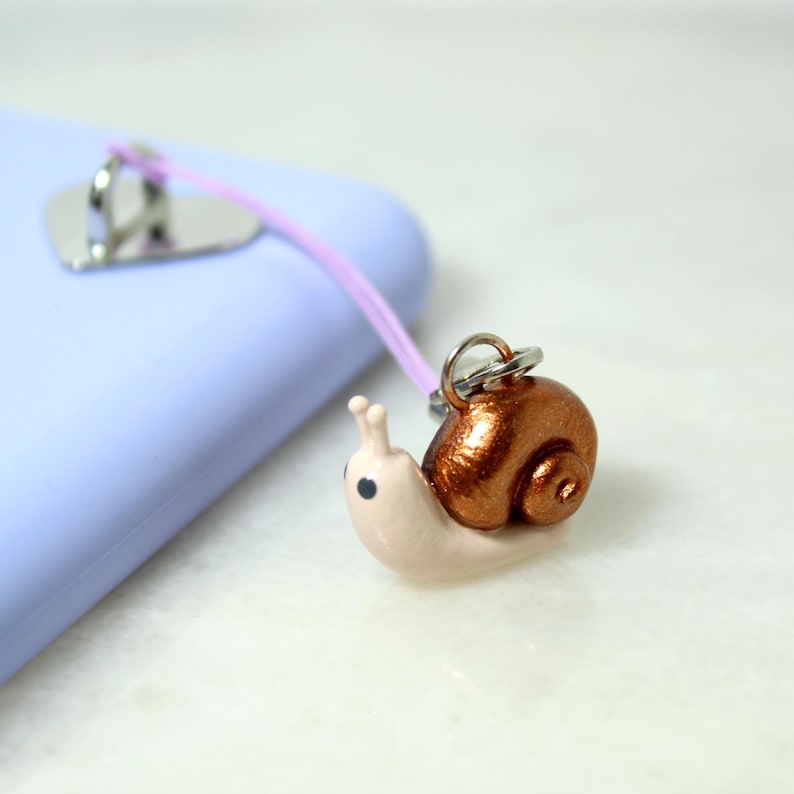 May include: A handmade snail phone charm. The snail has a metallic copper shell and a light pink body. It is attached to a lavender cord and a silver-colored phone grip.