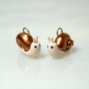 May include: Two handmade snail charms. Each charm features a light pink body, a metallic copper-colored shell, and black eyes. The snails have small antennae and a silver-colored loop for attaching to jewelry or other crafts.