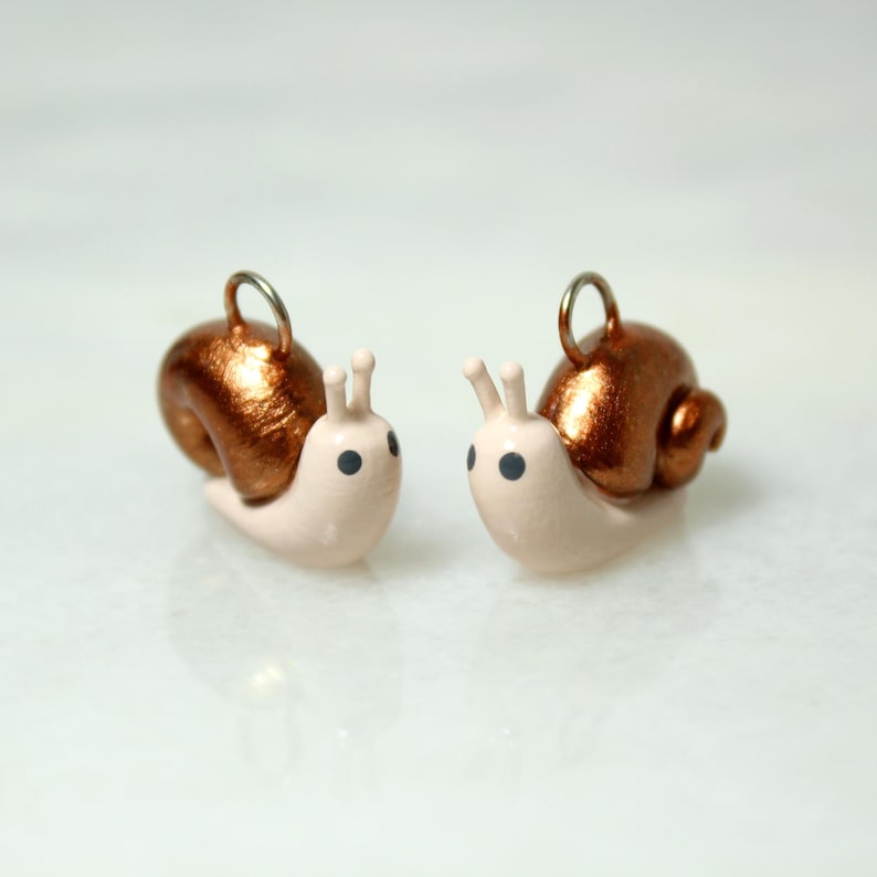May include: Two handmade snail charms. Each charm features a light pink body, a metallic copper-colored shell, and black eyes. The snails have small antennae and a silver-colored loop for attaching to jewelry or other crafts.