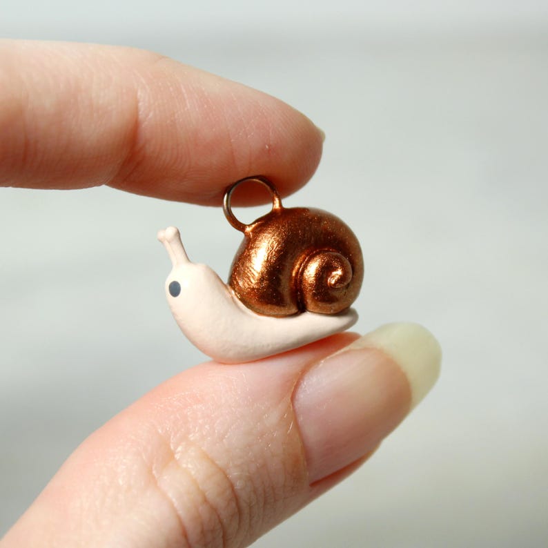 May include: A miniature snail charm with a metallic copper-colored shell and a cream-colored body. The snail has a small loop for attaching to a necklace or bracelet. The charm is held between two fingers.